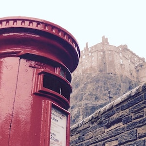 May include: A red vintage style post box with a sign that says "Mail Collection Times: Monday to Friday 9:30am, Saturday 2:15pm" in front of Edinburgh Castle.