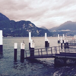 May include: A view of a lake with mountains in the background. A wooden dock with black and white poles extends into the water. The dock has a metal railing and a ramp leading to the shore.