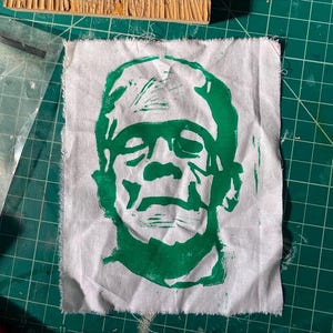 May include: Green and white fabric patch with a printed image of Frankenstein's face.