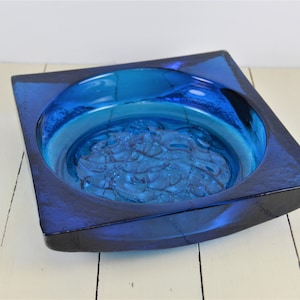 May include: A square blue glass ashtray with a raised, textured design in the center. The ashtray is made of thick, heavy glass and has a deep well.