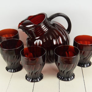 May include: A set of six ruby red glass tumblers and a matching pitcher. The glasses have a swirl design and the pitcher has a ribbed design.
