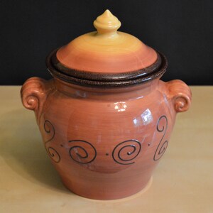 May include: A terracotta crock with a lid, featuring a brown rim and a swirl design. The crock has two handles and is decorated with a brown swirl pattern.