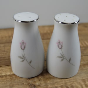 May include: A pair of white ceramic salt and pepper shakers with a silver rim and a pink rose design. The shakers have a classic shape and are likely part of a dining set. The rose is depicted in a delicate, watercolor style.