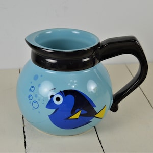 May include: A light blue ceramic mug with a black handle and rim. The mug features a cartoon image of a blue fish with yellow fins and the mug has a round shape. Bubbles are also depicted on the mug.