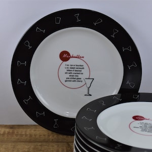 May include: A set of white ceramic dinner plates with black rims and a cocktail recipe printed on each plate. The recipe on the top plate is for a Manhattan cocktail. The recipe includes 3 oz. rye or bourbon, 1/2 oz. sweet vermouth, bitters if desired, stir with cracked ice, strain into a pre-chilled glass, and garnish with a cherry.
