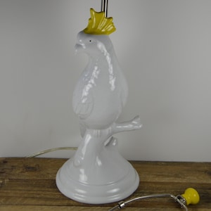 May include: White ceramic table lamp shaped like a cockatoo with a yellow crest. The lamp is on a wooden surface and has a yellow cord.