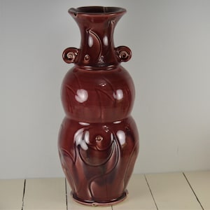 May include: A deep burgundy ceramic vase with a unique, bulbous shape and intricate, swirling designs. The vase has a wide mouth and two small handles on the sides.