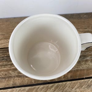 Puede incluir: Una taza de cer&aacute;mica blanca con un borde negro en el asa. La taza est&aacute; vac&iacute;a y se encuentra sobre una superficie de madera. El interior de la taza es de color blanco cremoso.