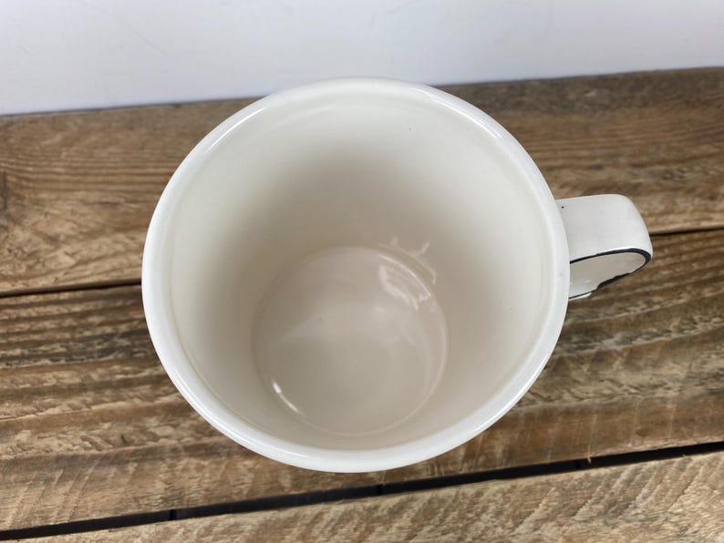 Puede incluir: Una taza de cer&aacute;mica blanca con un borde negro en el asa. La taza est&aacute; vac&iacute;a y se encuentra sobre una superficie de madera. El interior de la taza es de color blanco cremoso.