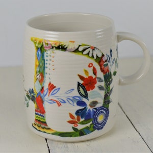 May include: Cream-colored ceramic mug with a large, colorful floral letter "B" design. The letter is filled with red, blue, and white flowers, green leaves, and other decorative elements. The mug has a curved handle.