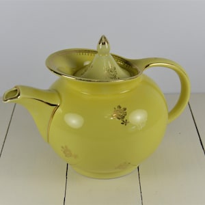 May include: A yellow teapot with a gold trim and floral design. The teapot has a lid with a knob on top.