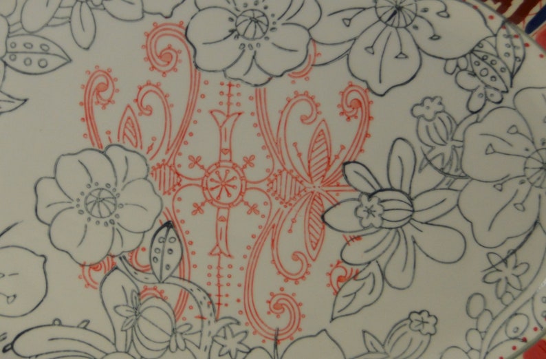 May include: A white ceramic plate with a black and red floral design. The design features a large red floral pattern in the center of the plate, surrounded by smaller black floral patterns.