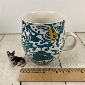 K&ouml;nnte beinhalten: Eine wei&szlig;e Keramiktasse mit einem t&uuml;rkis-wei&szlig;en Blumendesign und einem gelben Buchstaben "b". Neben der Tasse befindet sich eine kleine Corgi-Figur. Ein Holzlineal ist ebenfalls im Bild.