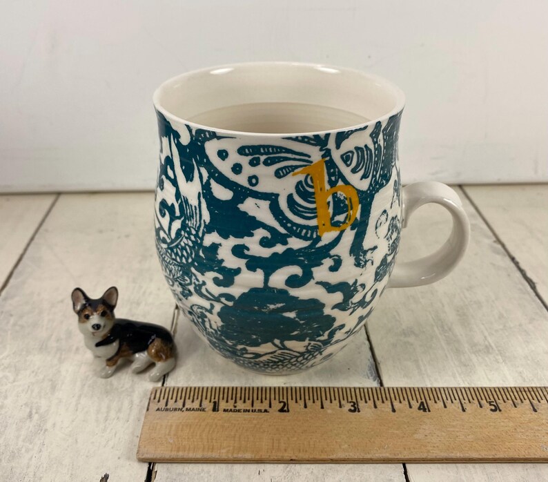 K&ouml;nnte beinhalten: Eine wei&szlig;e Keramiktasse mit einem t&uuml;rkis-wei&szlig;en Blumendesign und einem gelben Buchstaben "b". Neben der Tasse befindet sich eine kleine Corgi-Figur. Ein Holzlineal ist ebenfalls im Bild.