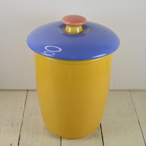 May include: A yellow ceramic canister with a blue lid and a pink knob. The canister has a smooth, glossy finish.