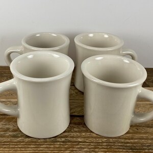 May include: Four off-white ceramic mugs with rounded bodies and curved handles are arranged on a wooden surface. The mugs have a simple, classic design, suitable for coffee or tea. The mugs are empty and ready to be used.