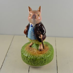 May include: A porcelain figurine of a pig wearing a blue jacket, yellow pants, and a brown hat. The pig is holding a walking stick and a brown bag. The figurine is standing on a green base with brown grass.