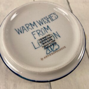 K&ouml;nnte beinhalten: Wei&szlig;e Keramikschale mit blauem Rand und dem Text "WARM WISHES FROM LONDON 2023" in blauer Schrift. Die Schale ist mit "Handwash Only" und "Made in China" von Anthropologie gekennzeichnet.