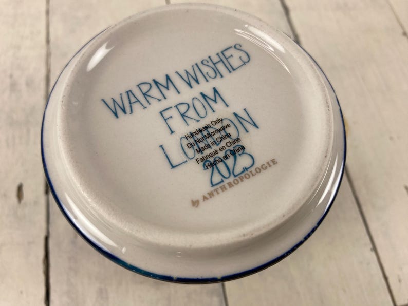K&ouml;nnte beinhalten: Wei&szlig;e Keramikschale mit blauem Rand und dem Text "WARM WISHES FROM LONDON 2023" in blauer Schrift. Die Schale ist mit "Handwash Only" und "Made in China" von Anthropologie gekennzeichnet.