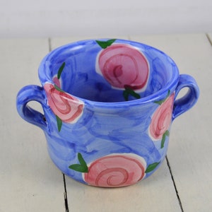 May include: A blue ceramic bowl with two handles and a floral design. The bowl is decorated with pink roses and green leaves.