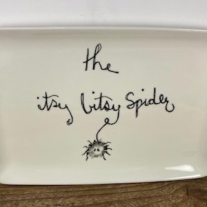 May include: A rectangular, off-white ceramic tray with rounded corners. The words "the itsy bitsy Spider" are written in black script, with a small spider illustration below. The tray is on a wooden surface.