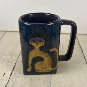 May include: A navy blue ceramic mug with a square shape and a black handle. The mug features a tan cat design with blue accents. The mug is a unique piece of kitchenware.