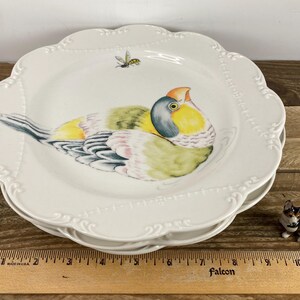 May include: A stack of white ceramic plates with a scalloped edge and a painted bird design. The bird features yellow, green, and blue plumage, with an orange beak. A small bee is painted near the bird. A miniature dog figurine is also present.