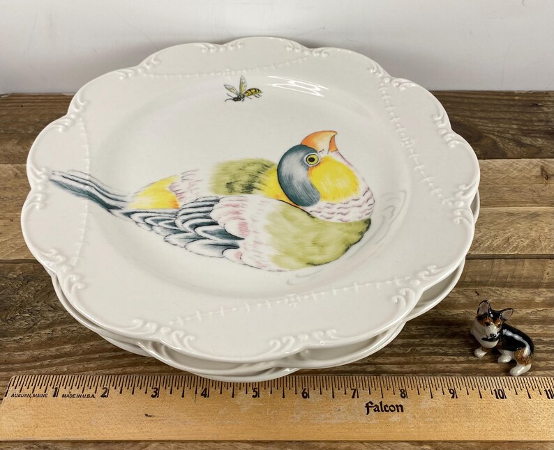 May include: A stack of white ceramic plates with a scalloped edge and a painted bird design. The bird features yellow, green, and blue plumage, with an orange beak. A small bee is painted near the bird. A miniature dog figurine is also present.