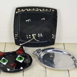 May include: Three ceramic plates, one black with a white cat face design, one black with a red and green cat face design, and one silver oval plate with decorative handles.