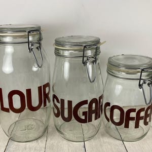 May include: Three clear glass jars with metal hinged lids. The jars are labeled in dark red lettering: "FLOUR", "SUGAR", and "COFFEE". The jars are different sizes, suitable for kitchen storage.