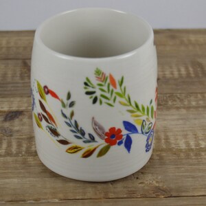 May include: A white ceramic mug with a floral design. The colorful design includes red, blue, yellow, and green flowers and leaves. The mug is cylindrical and sits on a wooden surface.