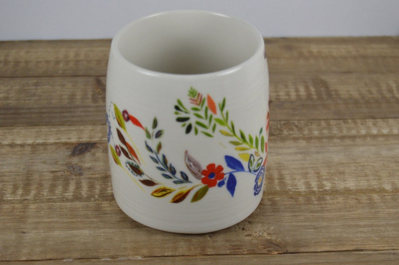 May include: A white ceramic mug with a floral design. The colorful design includes red, blue, yellow, and green flowers and leaves. The mug is cylindrical and sits on a wooden surface.