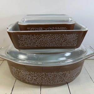 May include: A set of two brown glass baking dishes with white floral line art patterns. The rectangular dish features a clear glass lid, while the larger oval dish also includes a matching clear glass lid. Both pieces have integrated handles.