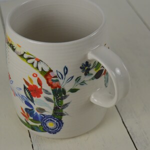 May include: Cream-colored ceramic mug with a curved handle and a floral design. The flowers are in shades of red, blue, white, and yellow, with green leaves. The mug is on a white wooden surface.