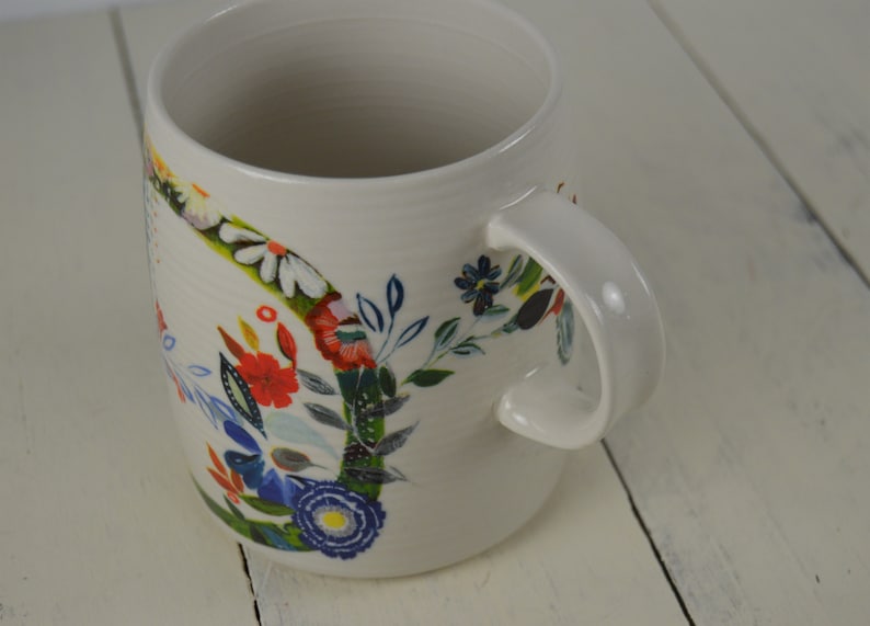 May include: Cream-colored ceramic mug with a curved handle and a floral design. The flowers are in shades of red, blue, white, and yellow, with green leaves. The mug is on a white wooden surface.