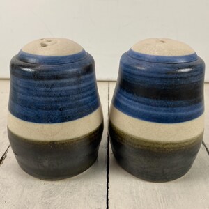 May include: A pair of ceramic salt and pepper shakers. Each shaker is cylindrical with a light beige top, blue and black bands, and a dark base. The shakers have a rustic, handmade appearance.