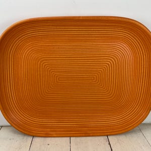 May include: An orange rectangular serving tray with a textured, concentric design. The tray has rounded corners and a slightly raised edge. The surface is covered in parallel lines that create a ribbed effect. The tray is sitting on a white wooden surface.