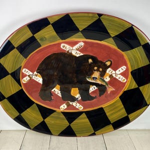 May include: Oval serving platter with a black and yellow checkered border. The center features a brown bear holding a fish, set against a red oval background. Decorative birch logs surround the bear.