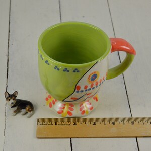 May include: A green and white ceramic mug with a colorful floral design and a red handle. The mug has a rounded base and a wide, open top. A small ceramic figurine of a dog is in the foreground.