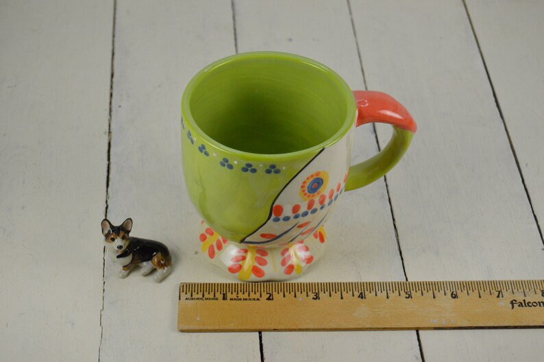 May include: A green and white ceramic mug with a colorful floral design and a red handle. The mug has a rounded base and a wide, open top. A small ceramic figurine of a dog is in the foreground.