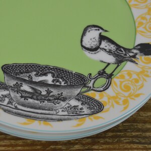 K&ouml;nnte beinhalten: Ein gr&uuml;ner und wei&szlig;er Keramikteller mit gelbem Blumendesign. Eine Schwarzwei&szlig;illustration eines Vogels und einer Teetasse befinden sich auf dem Teller.