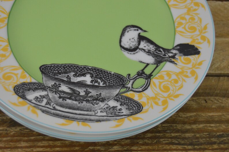 K&ouml;nnte beinhalten: Ein gr&uuml;ner und wei&szlig;er Keramikteller mit gelbem Blumendesign. Eine Schwarzwei&szlig;illustration eines Vogels und einer Teetasse befinden sich auf dem Teller.