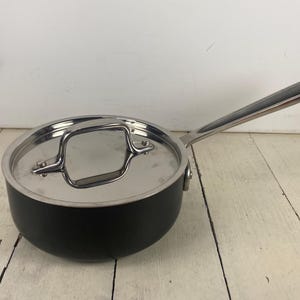 May include: A black saucepan with a matte finish and a polished stainless steel lid. The pan features a long, slender stainless steel handle extending from the side and a matching metal loop handle centered on the circular lid.