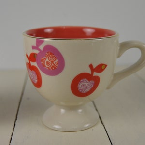 May include: Two white ceramic mugs with a footed base. The mugs have a red rim and are decorated with pink and red apples with a gold outline.