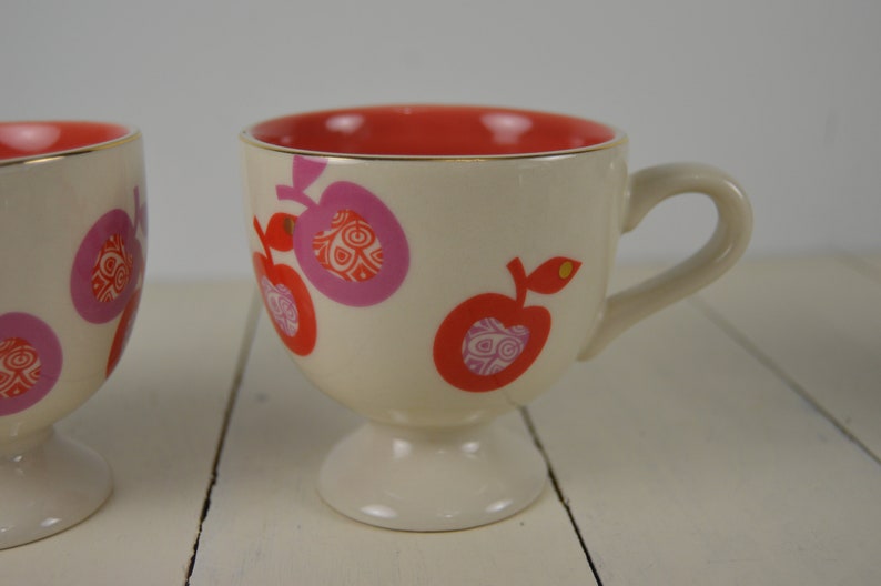 May include: Two white ceramic mugs with a footed base. The mugs have a red rim and are decorated with pink and red apples with a gold outline.