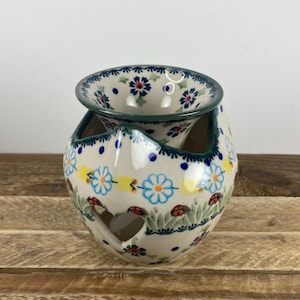 May include: A ceramic garlic keeper with a floral design. The off-white keeper features blue, yellow, and red flowers, green grass, and ladybugs. The rim and interior are green. The keeper has a heart-shaped opening.