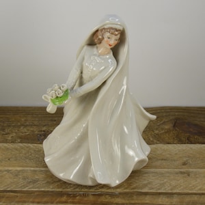 May include: A porcelain figurine of a bride wearing a white gown and veil, holding a bouquet of white flowers.