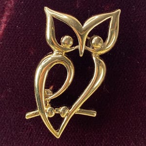 Op de afbeelding: Een goudkleurige uilbroche. De broche heeft een gestileerd uilontwerp met grote ogen en een gebogen lichaam. De uil zit op een tak. De broche is gemaakt van glanzend, reflecterend metaal.
