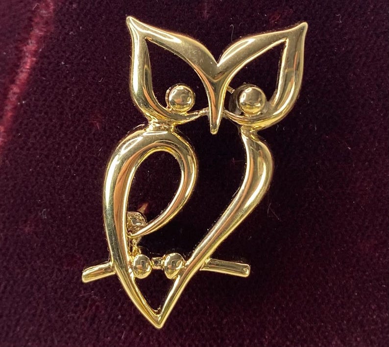 Op de afbeelding: Een goudkleurige uilbroche. De broche heeft een gestileerd uilontwerp met grote ogen en een gebogen lichaam. De uil zit op een tak. De broche is gemaakt van glanzend, reflecterend metaal.
