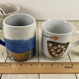K&ouml;nnte beinhalten: Zwei Keramikbecher mit einzigartigen Designs. Ein Becher hat ein blau-wei&szlig;es Design mit goldfarbenem Boden. Der andere Becher ist wei&szlig; mit einem braunen und rosa Design. Eine kleine Hundefigur ist ebenfalls vorhanden.